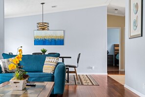 Een woonkamer met een blauwe bank, een bijzettafeltje met een bloemenvaas, een hanglamp en een schilderij aan de muur.