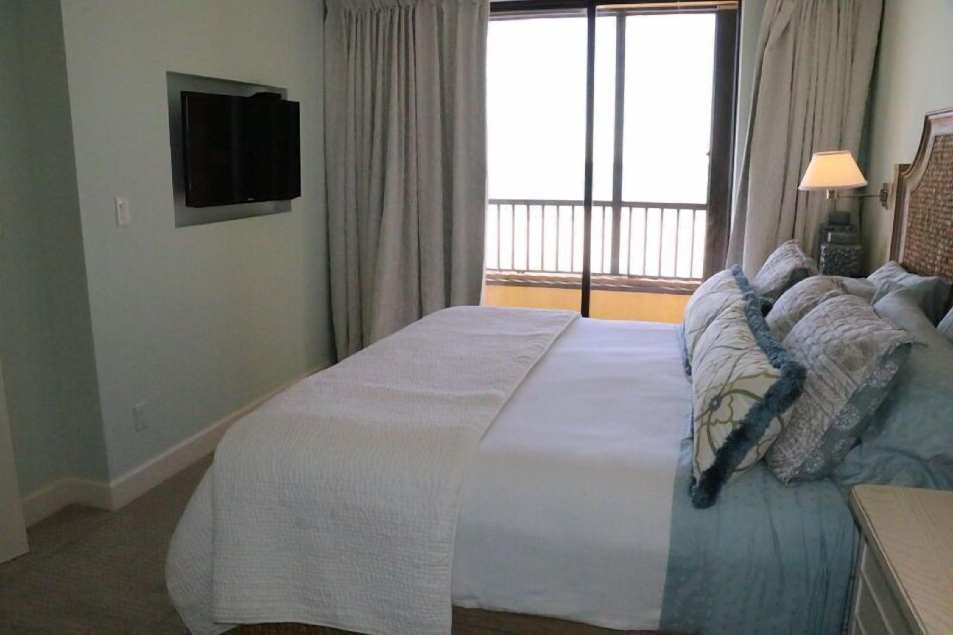 A hotel room with a large bed, a flat-screen TV mounted on the wall, a balcony with a railing, and a nightstand with a lamp.
