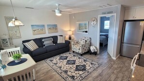 A modern kitchen with a refrigerator, stove, and dining area. There is a sofa and a chair in the living area, and artwork on the walls. - Gulf Holiday Unit 12- Close to Beach and Siesta Key Village! (Siesta Key)