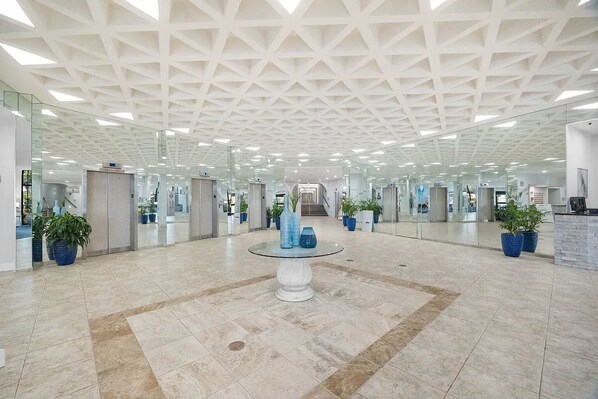 En rummelig lobby med et geometrisk loftsdesign, glasvægge og et rundt bord i midten med dekorative genstande.