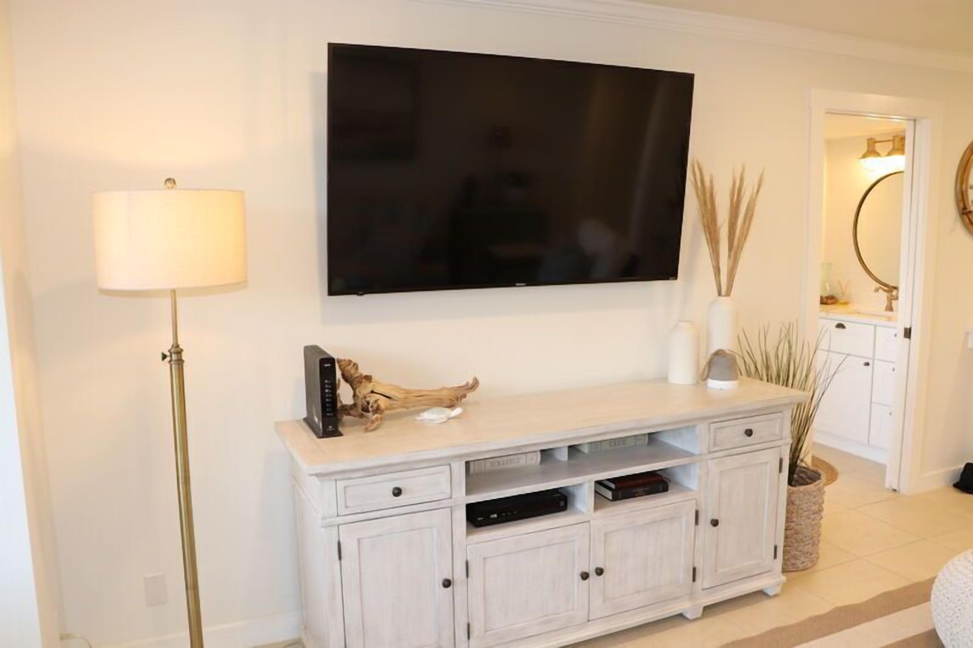 Een woonkamer met een flatscreen televisie aan de muur, een wit bergkastje met lades, een vloerlamp en een kleine potplant.