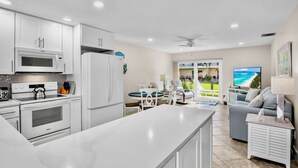 A modern kitchen with white cabinets, a microwave, stove, and refrigerator. There is a dining area with a table and chairs, and a living room with a couch and TV. - Jamaica Royale 1 UPDATED & CLOSEST TO BEACH (Siesta Key)