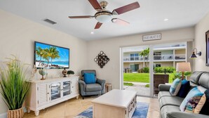 A living room with a ceiling fan, a TV displaying a beach scene, a couch with decorative pillows, and a view of a balcony with greenery. - Perfect Beachfront Couples Retreat at Jamaica Royale 002 in Siesta Key! (Siesta Key)