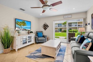 Un salon doté d’un ventilateur de plafond, d’un téléviseur diffusant une scène de plage, d’un canapé agrémenté de coussins décoratifs et offrant une vue sur un balcon verdoyant.