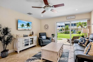 A living room with a ceiling fan, a TV, a couch, a coffee table, a chair, and a view of a building labeled 'BEACH' through a glass door.