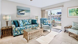 Ein Wohnzimmer mit einer Couch, einem Sessel und einem Glastisch. Es gibt einen Deckenventilator, ein Gemälde eines Bootes und einen Blick auf einen Pool und Gebäude im Freien.