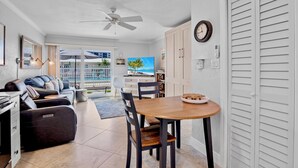 A modern living room with a sofa, dining table, and chairs. There is a TV, a ceiling fan, and a view of the outside. - Jamaica Royale 009 Updated and across from pool! (Siesta Key)