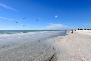 Ein Strand mit blauem Himmel, ruhigem Wasser und wenigen Personen, die am Ufer entlanggehen.