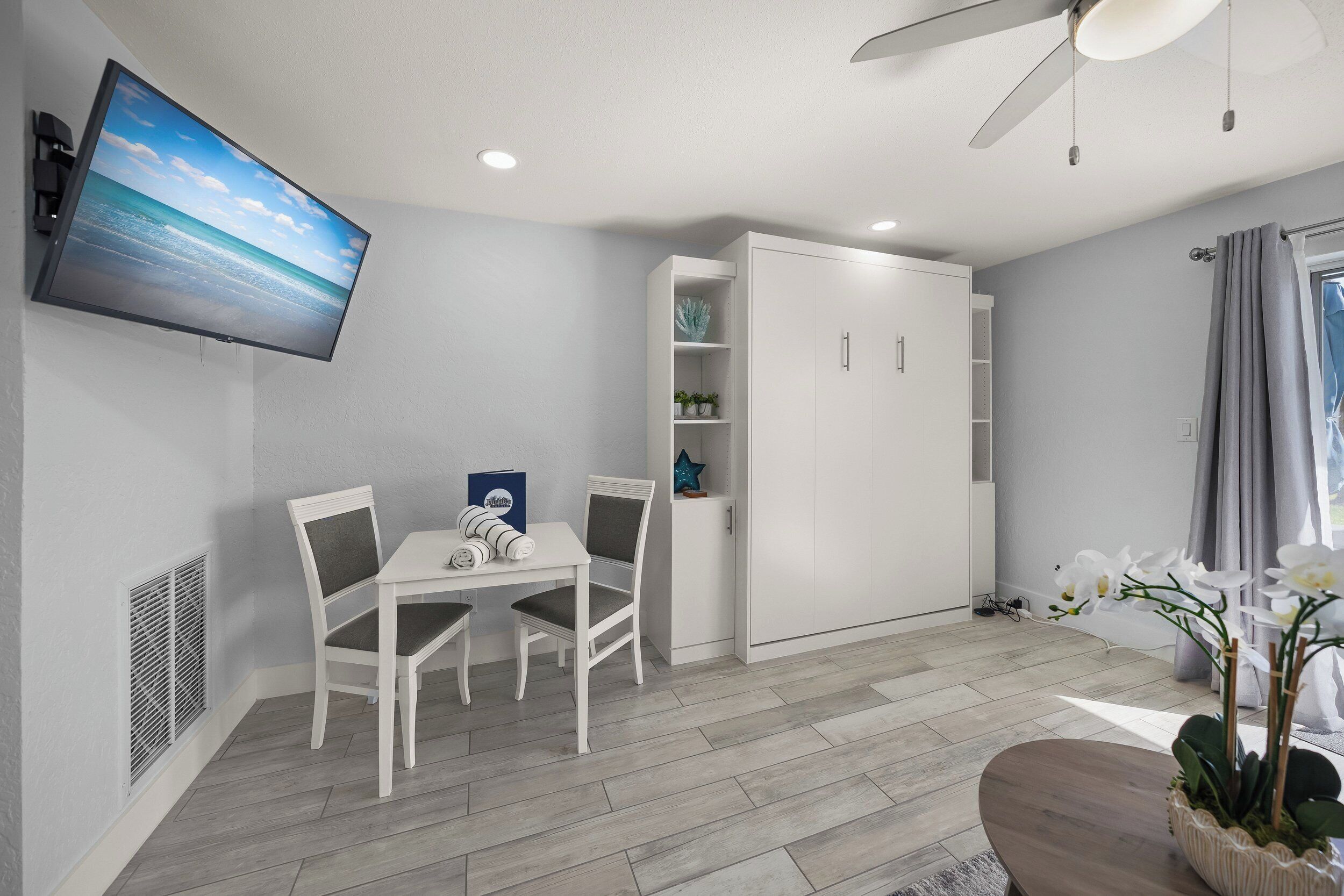 A modern living room with a flat-screen TV mounted on the wall, a dining area with a table and chairs, a white wardrobe, and a ceiling fan.
