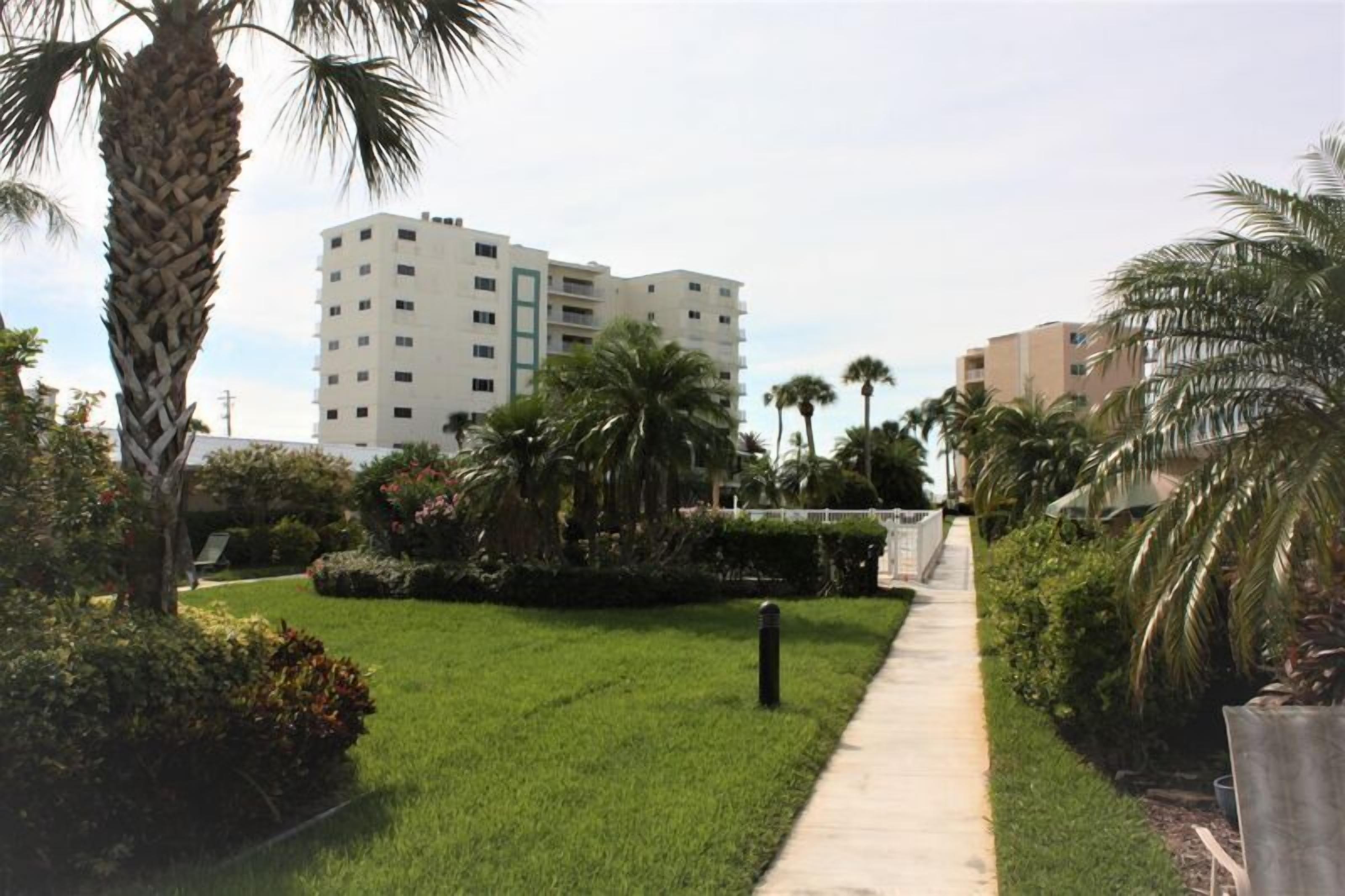 A pathway leading to a building with a modern design, surrounded by well-maintained greenery and palm trees.