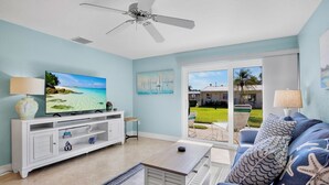 A living room with a TV, white entertainment unit, blue sofa, and a view of a green lawn and houses through the glass door. - Jamaica Royale 022 Short walk to the beach! (Siesta Key)
