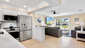 A modern kitchen with stainless steel appliances, a dining area with a gray sofa, and a view of a tropical landscape through the glass door. - Jamaica Royale 028 Washer and Dryer Plus Half Bath (Siesta Key)