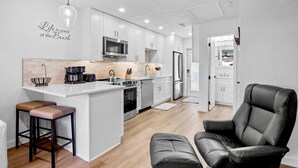 A modern kitchen with white cabinets, a central island with stools, a black leather chair, and a microwave. - Jamaica Royale 031 Nicely Updated! (Siesta Key)