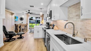 A modern kitchen with white cabinets, a stainless steel sink, and a dining area with a view of the outdoors. - Jamaica Royale 031 Nicely Updated! (Siesta Key)