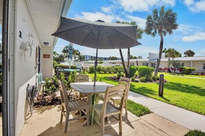 An outdoor patio with a table and chairs, a large umbrella, and a view of a well-maintained lawn and buildings in the background.