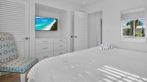 A bedroom with a large bed, a TV mounted on the wall, a window with a view of palm trees, and a patterned chair. - Jamaica Royale 033 Close to pool! (Siesta Key)