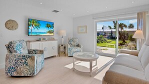 A living room with a beach-themed painting, a white sofa, two patterned armchairs, a small round table, and a view of the outdoors through large windows. - Jamaica Royale 033 Close to pool! (Siesta Key)