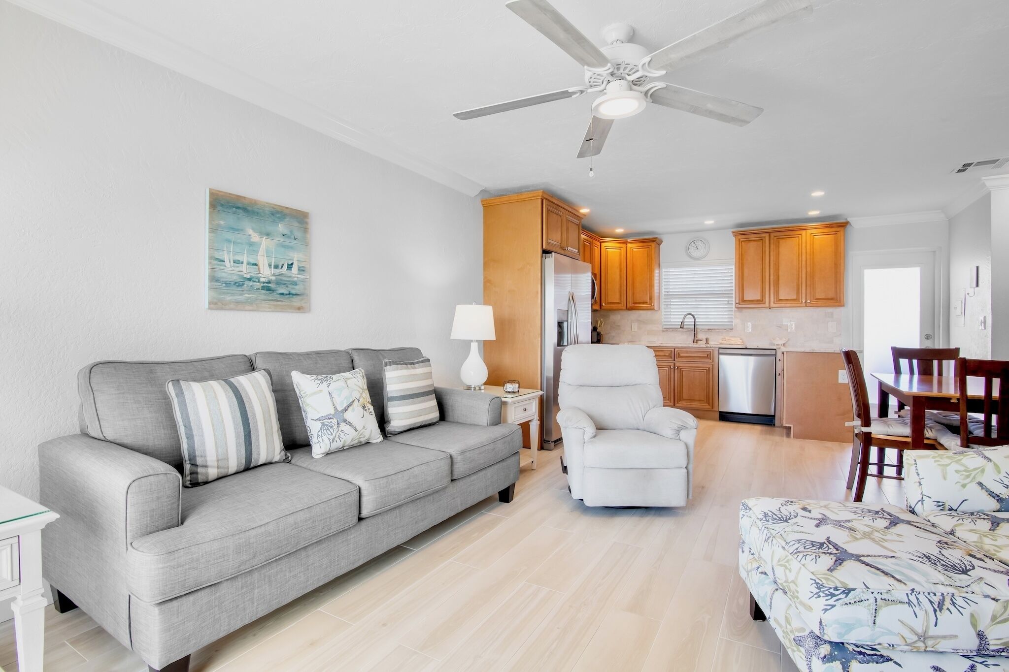 A living room with a sofa, armchair, and a dining table with chairs. There is a kitchen area with cabinets and a refrigerator.