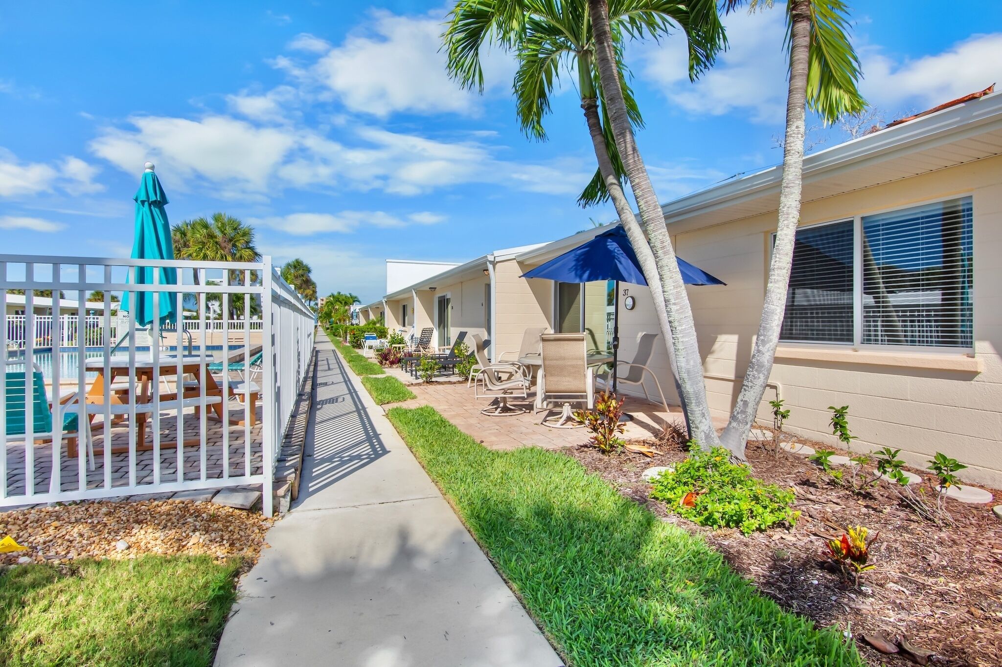 A residential area with a paved walkway, a white fence, and a small patio with outdoor furniture.