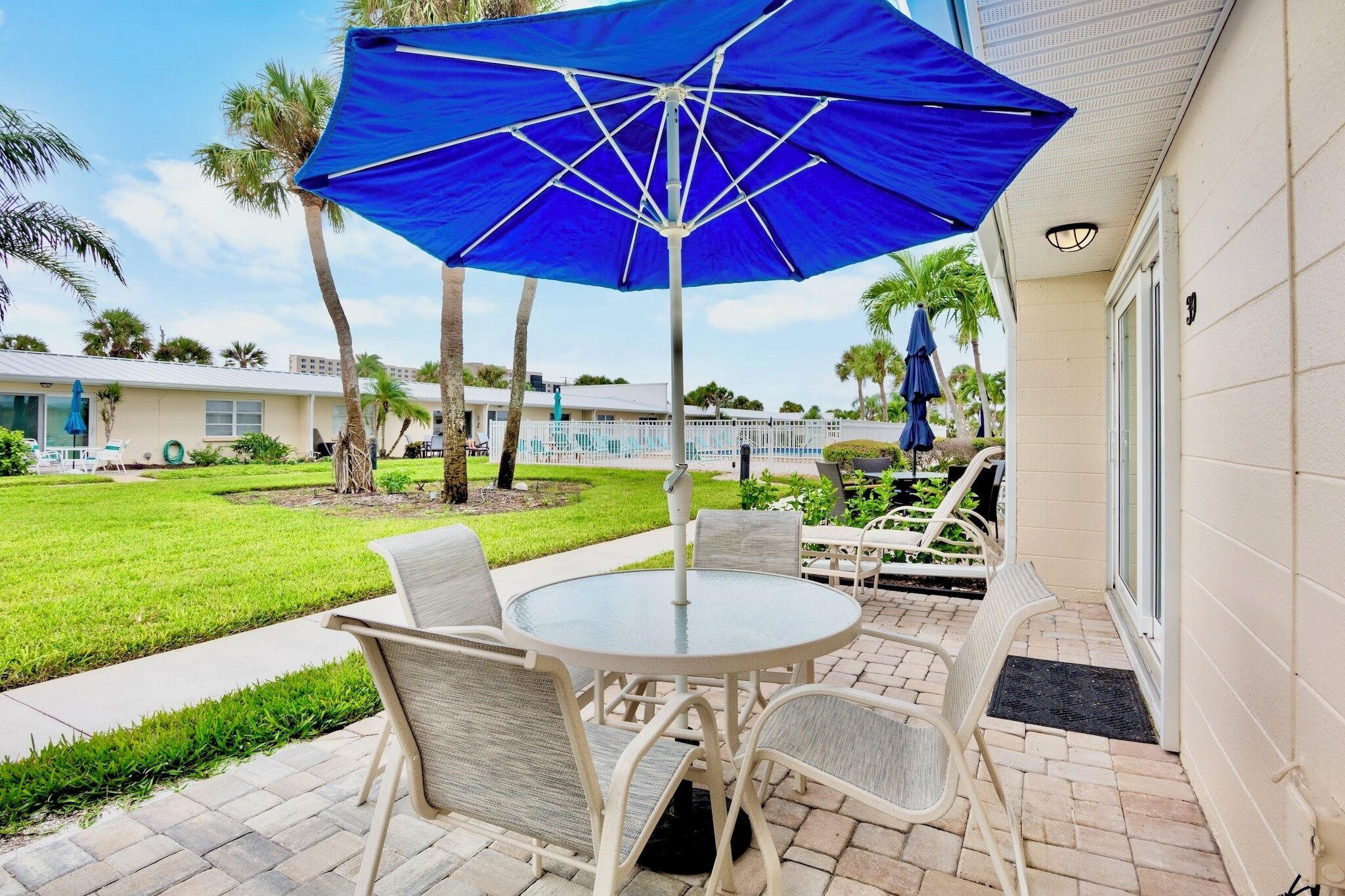 A patio with a round table and chairs, a blue umbrella, and a pool in the background.