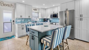 A modern kitchen with white cabinets, a central island with striped chairs, a stainless steel refrigerator, and a backsplash with blue tiles. - Jamaica Royale 048 Updated and close to the pool! (Siesta Key)