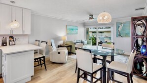 Cozinha moderna com armários brancos, área de jantar com mesa de vidro e sala de estar com sofá e abajur.