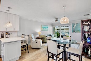 A modern kitchen with white cabinetry, a dining area with a glass table, and a living room with a sofa and a lamp.