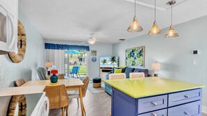 Une cuisine moderne avec un îlot central jaune, un coin repas avec des chaises en osier et un salon avec un canapé et des coussins bleus.