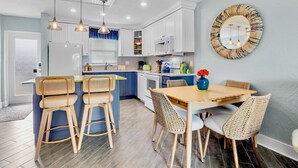 Une cuisine moderne avec des meubles blancs, un îlot central en bois doté de tabourets, et une table à manger avec des chaises en osier.