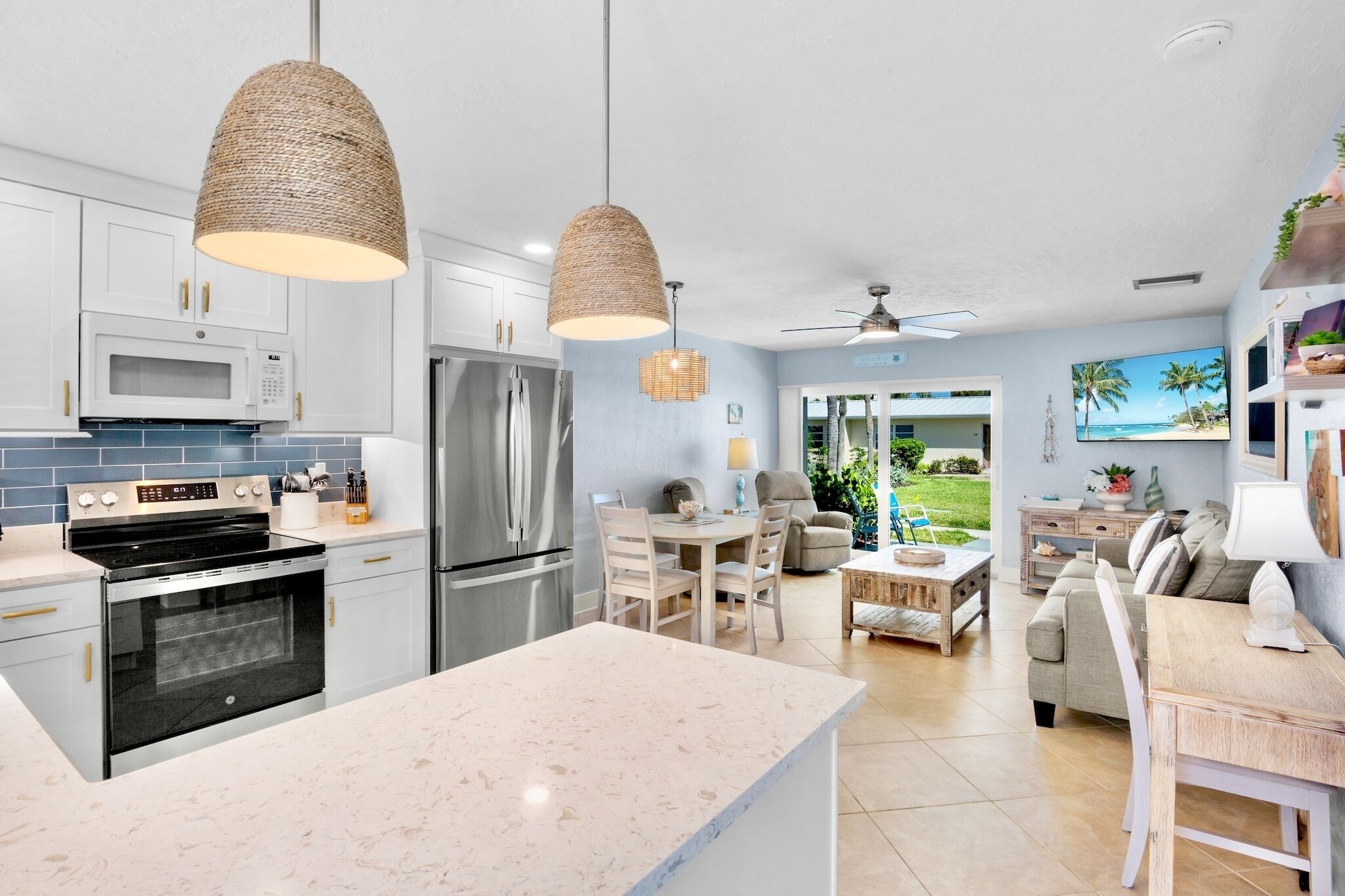 A modern kitchen with white cabinets, stainless steel appliances, and a large island. There is a dining area with a table and chairs, and a living room with a sofa and a TV.