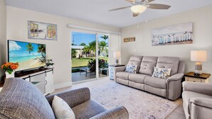 A living room with a sofa, a television, and a ceiling fan. There is a window with a view of palm trees and a beach. - Jamaica Royale 063 Great central location! (Siesta Key)