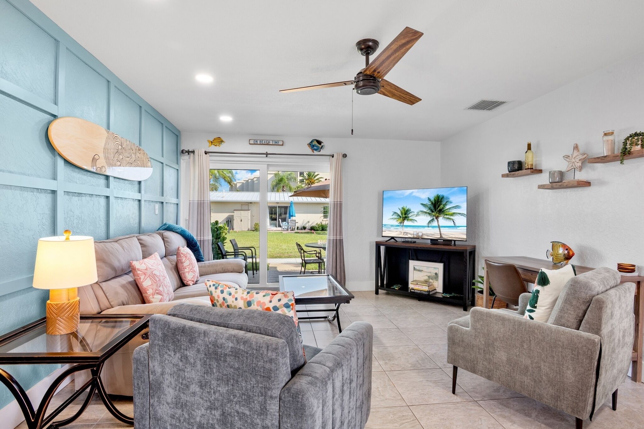 A living room with a sofa, armchairs, a coffee table, a TV, a ceiling fan, and a view of the outdoors.