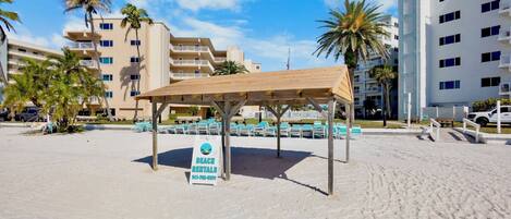 A beach rental area with a covered seating area, a sign for beach rentals, and buildings in the background.
