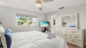 Un dormitorio con cama, ventilador de techo, televisor, cómoda y ventana con vista a palmeras.