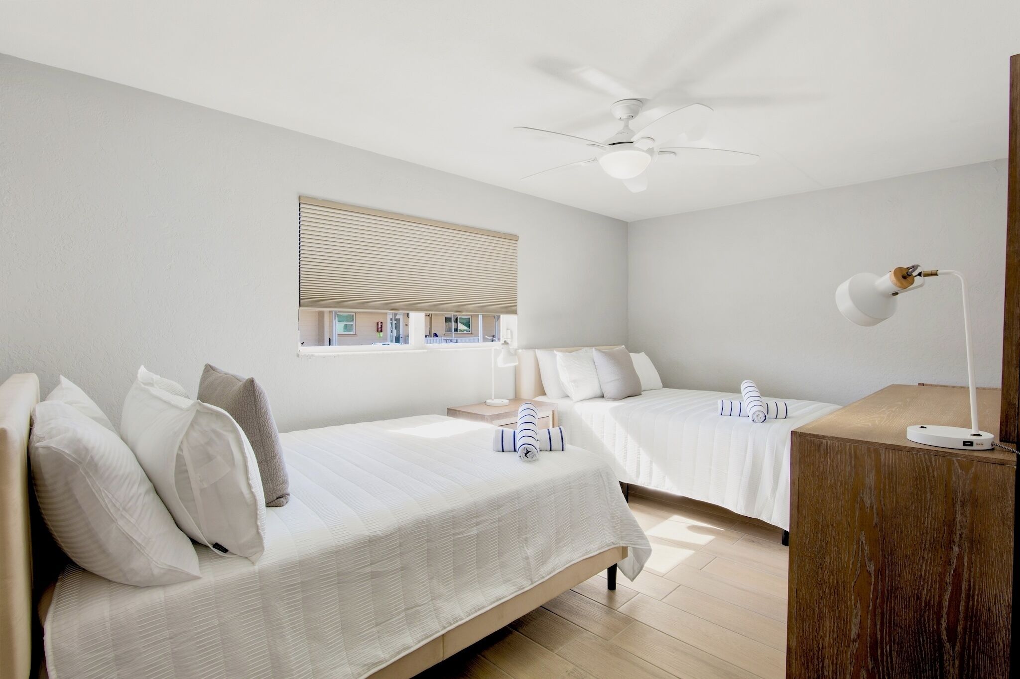 A hotel room with two beds, a ceiling fan, a desk with a lamp, and a window with blinds.