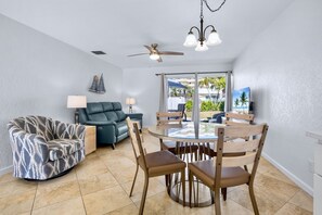 A living room with a sofa, a dining table with chairs, a ceiling fan, and a view of the outdoors.