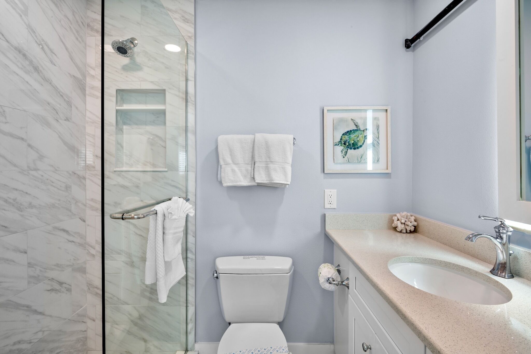 A bathroom with a glass shower enclosure, a toilet, a sink with a mirror, and a framed artwork on the wall.