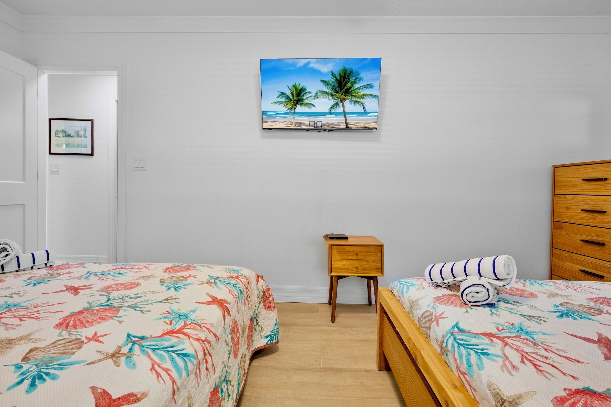 Une chambre avec deux lits, une commode en bois, une petite table de chevet et un tableau représentant une scène de plage.