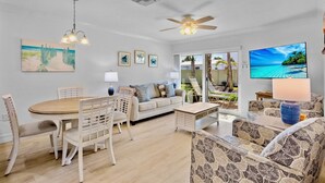 Un salon spacieux avec une table à manger en bois, des chaises blanches, un canapé et un téléviseur. On y trouve également un ventilateur de plafond, une grande fenêtre donnant sur l’extérieur et un luminaire au plafond.