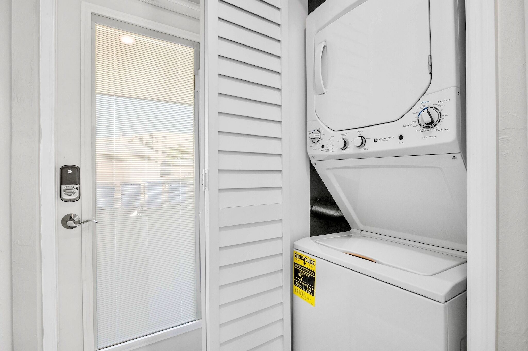 Une buanderie avec une porte blanche, une fenêtre avec des stores et un lave-linge muni d’une étiquette « Guide énergétique ».