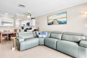 A modern living room with a sectional sofa, a dining area with a wooden table and chairs, a kitchen with white cabinets, and a painting of a pier on the wall.
