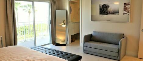 A bedroom with a bed, a grey armchair, a bench, a large mirror, and a framed picture on the wall.
