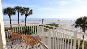 Een balkon met rieten stoelen en een tafel, met uitzicht op een strand met palmbomen en mensen.