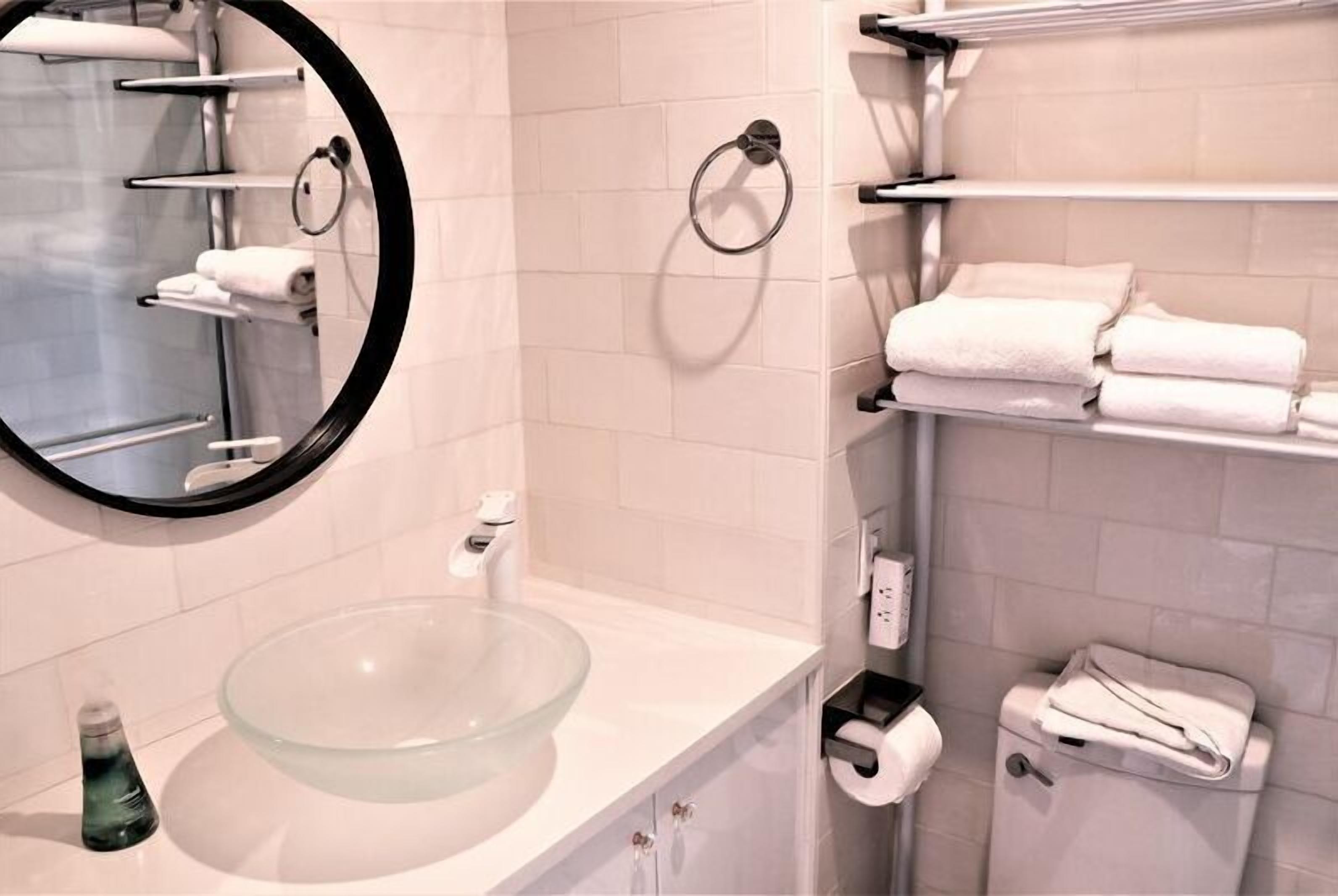 Un bagno moderno con un lavabo in vetro, uno specchio rotondo e pareti rivestite di piastrelle bianche.