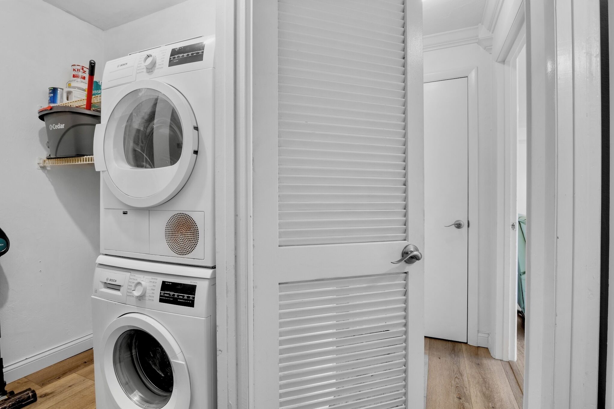 Une buanderie avec des machines à laver superposées, une porte à panneaux ajourés et une étagère contenant des produits de nettoyage.