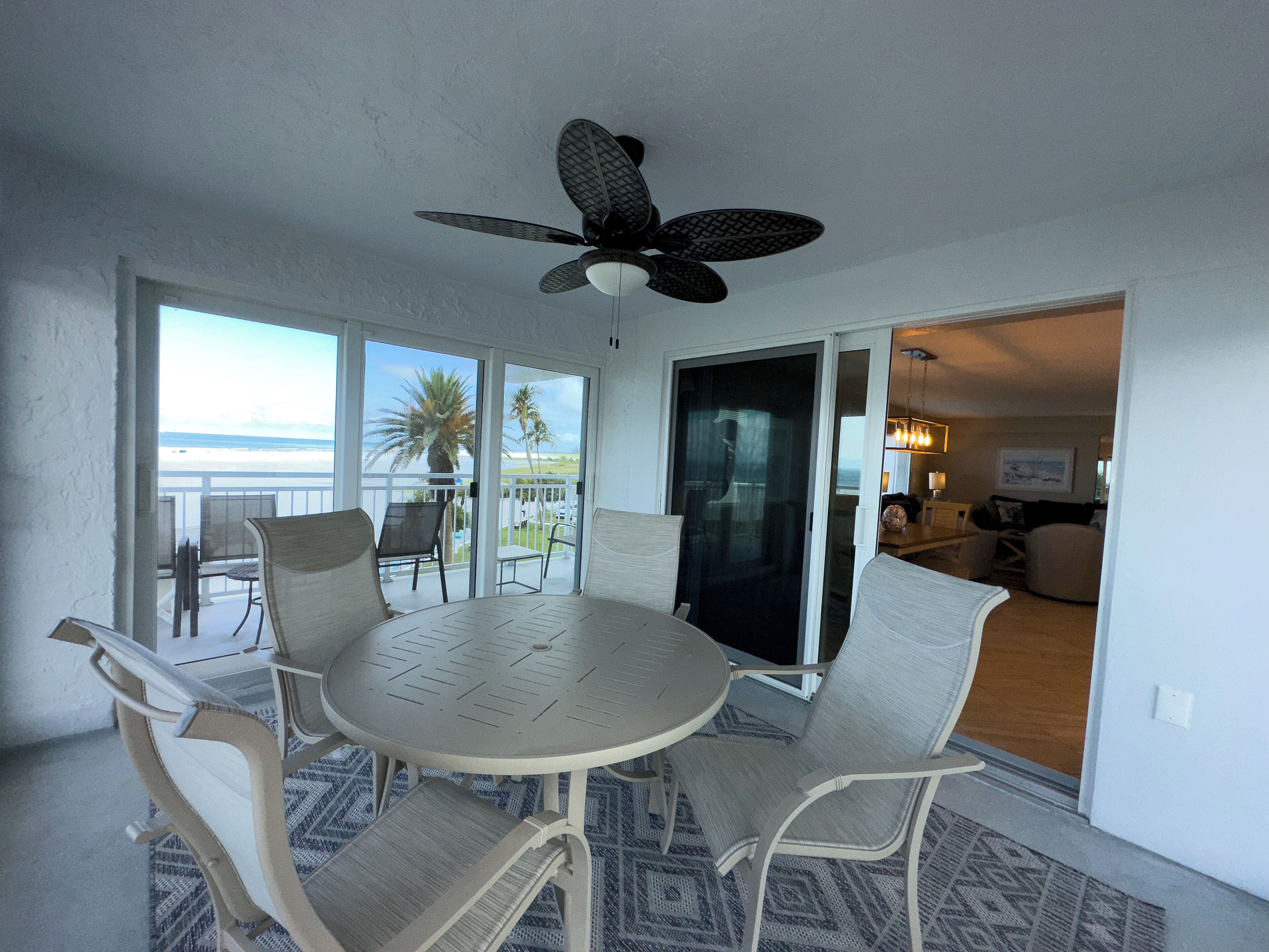 Ein Essbereich mit einem runden Tisch und Stühlen, einem Deckenventilator und großen Schiebeglasüren, die auf einen Balkon mit Blick auf den Strand und Palmen führen.