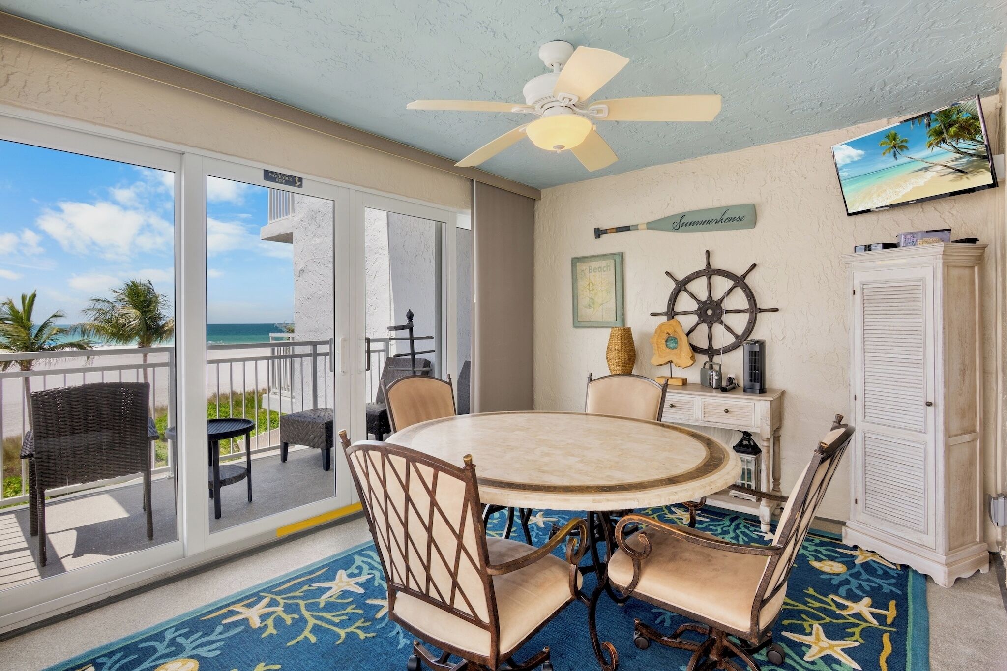 A room with a round table, chairs, a ceiling fan, a TV, a ship's wheel, and a view of the beach.