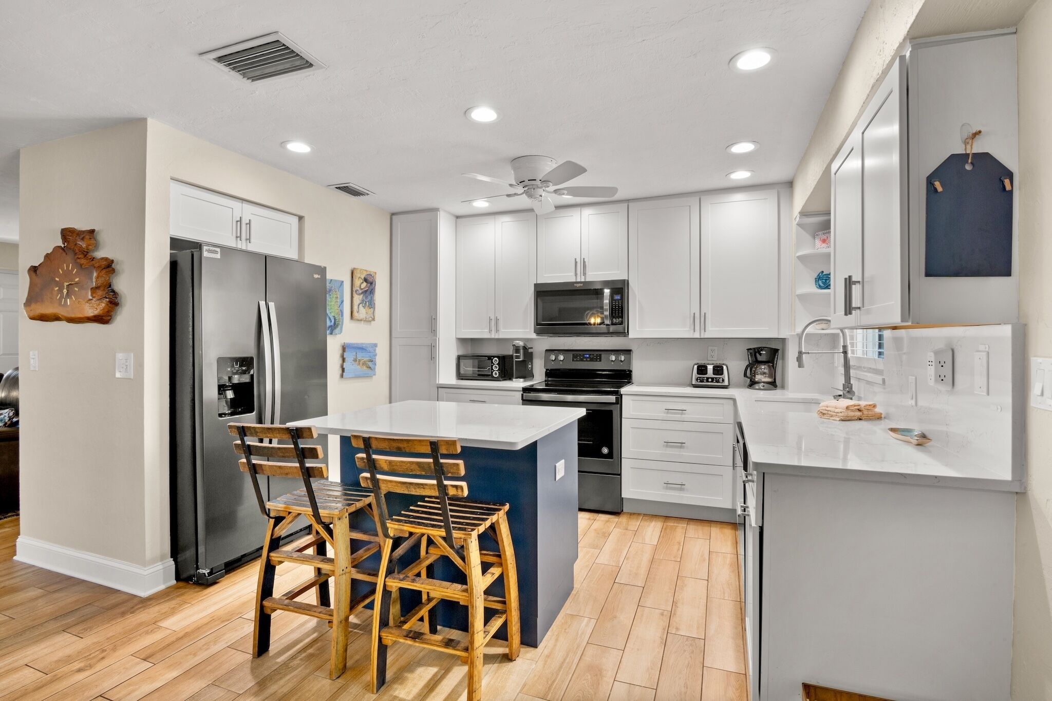 Una cocina moderna con armarios blancos, electrodomésticos de acero inoxidable, una isla central con taburetes y un ventilador de techo.