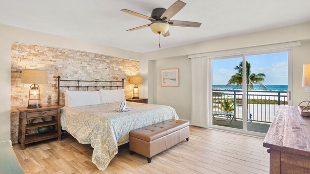 Una camera da letto con un letto, lampade sui comodini, una panca in legno, una ventola a soffitto e un balcone con vista sulla spiaggia.
