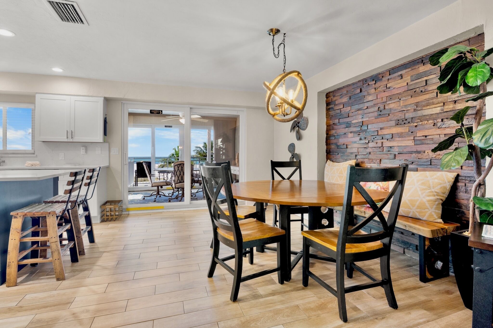 A modern kitchen with white cabinets, a wooden dining table, and chairs. There is a view of the outdoors through a glass door.